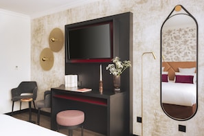 A modern hotel room with a flat-screen TV mounted on a dark wall, a round mirror with a decorative frame, a small desk with a chair, and a bedside table with a vase of flowers. - Appart'City Collection Genève Aéroport – Ferney Voltaire (Ferney-Voltaire)