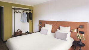 A hotel room with a large bed, a bedside table with a lamp, a wall-mounted TV, and a coat rack with hanging clothes. - Appart'City Collection Genève Aéroport – Ferney Voltaire (Ferney-Voltaire)
