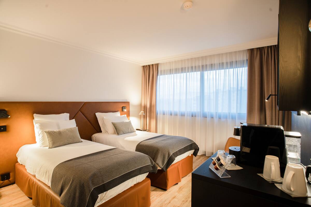 Une chambre d’hôtel avec deux lits, un bureau avec une machine à café et une grande fenêtre avec des rideaux.