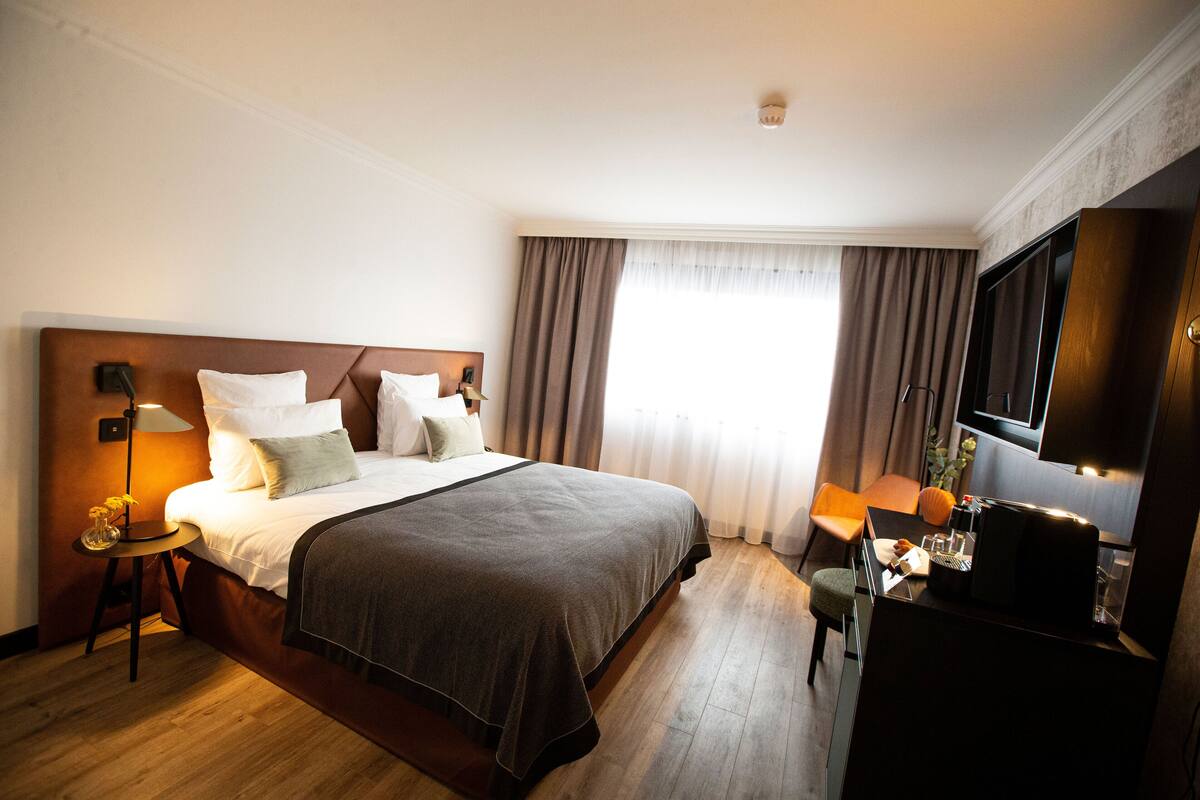 Une chambre d’hôtel avec un grand lit, un bureau avec une chaise, une télévision et une fenêtre avec des rideaux.