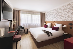 A hotel room with a bed, a desk, a chair, a TV, and a window with curtains.
