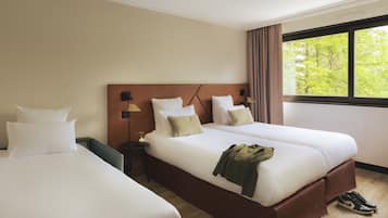 Een hotelkamer met twee bedden, een nachtkastje, een lamp en een raam met uitzicht op bomen.