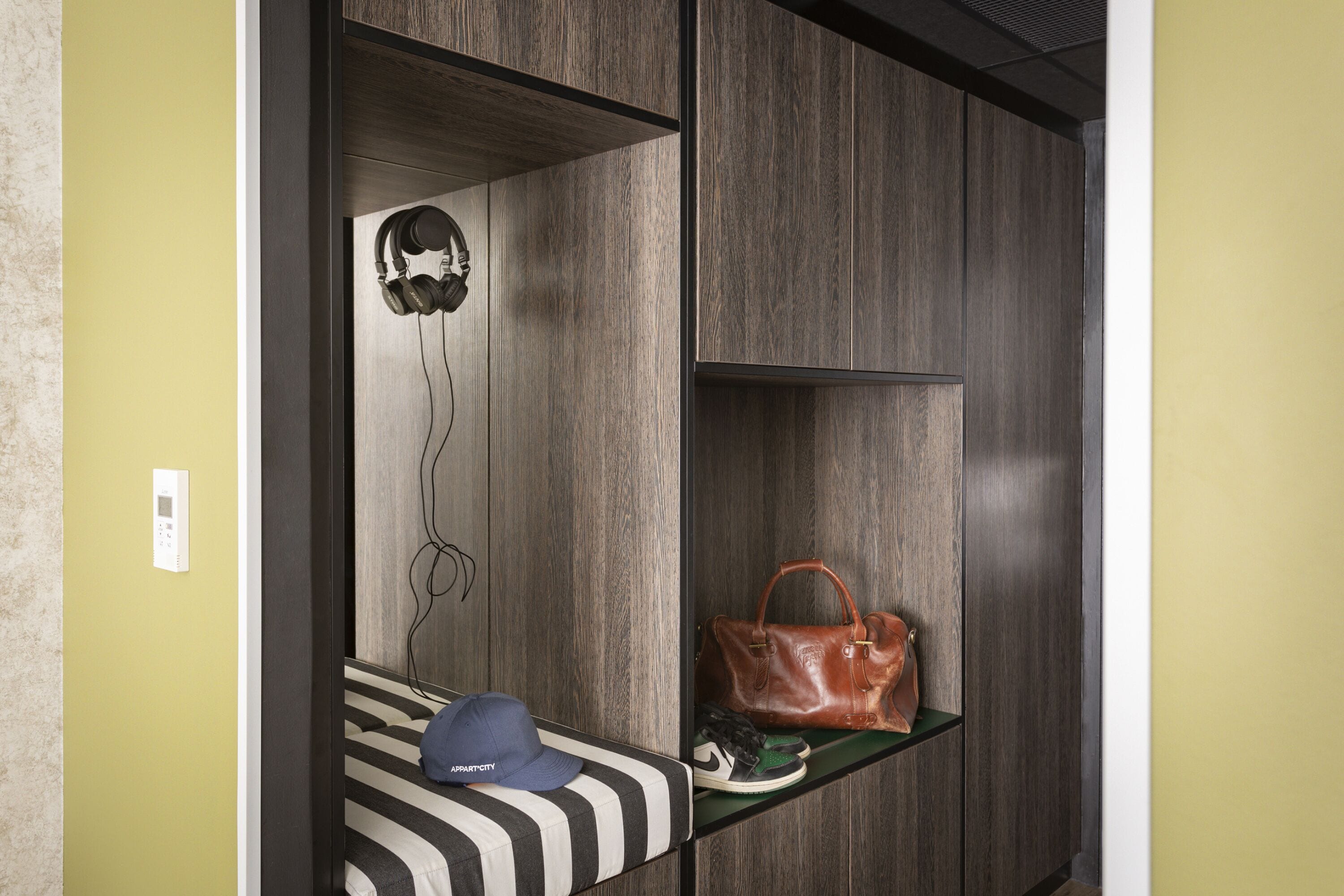 A room with a bed, a shelf with a handbag and shoes, and a wall-mounted thermostat.