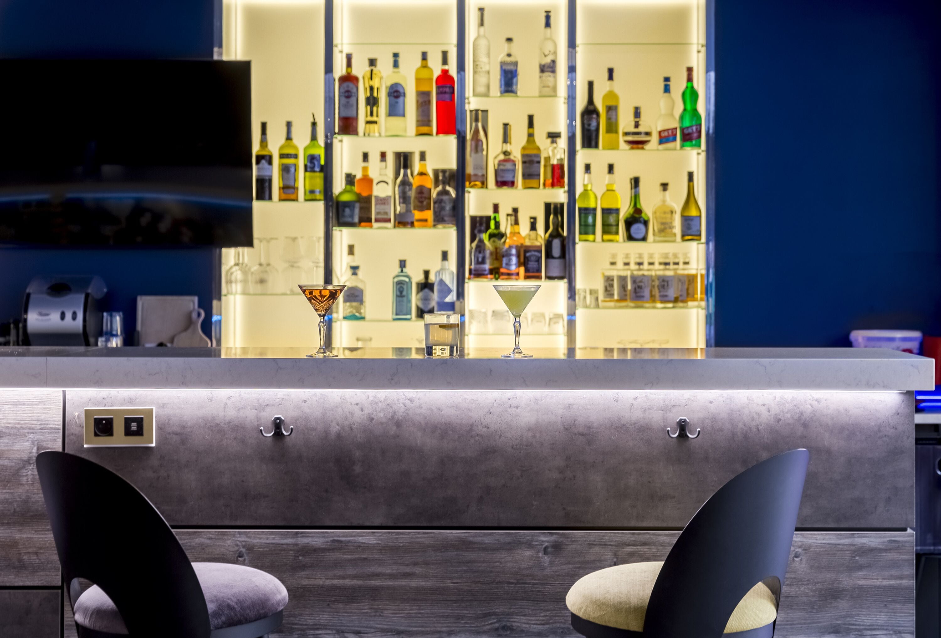 Zona de bar con mostrador de mármol, dos taburetes tapizados y varias botellas de licores en estantes al fondo.