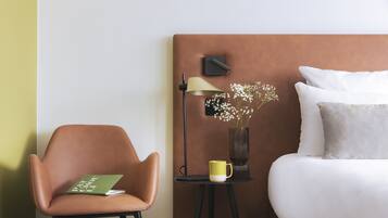 Hotelkamer met een bed, een stoel, een nachtkastje met lamp, een vaas met bloemen en een boek.