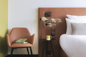 A hotel room with a bed, a chair, a nightstand with a lamp, a vase with flowers, and a book. - Appart'City Collection Genève Aéroport – Ferney Voltaire (Ferney-Voltaire)