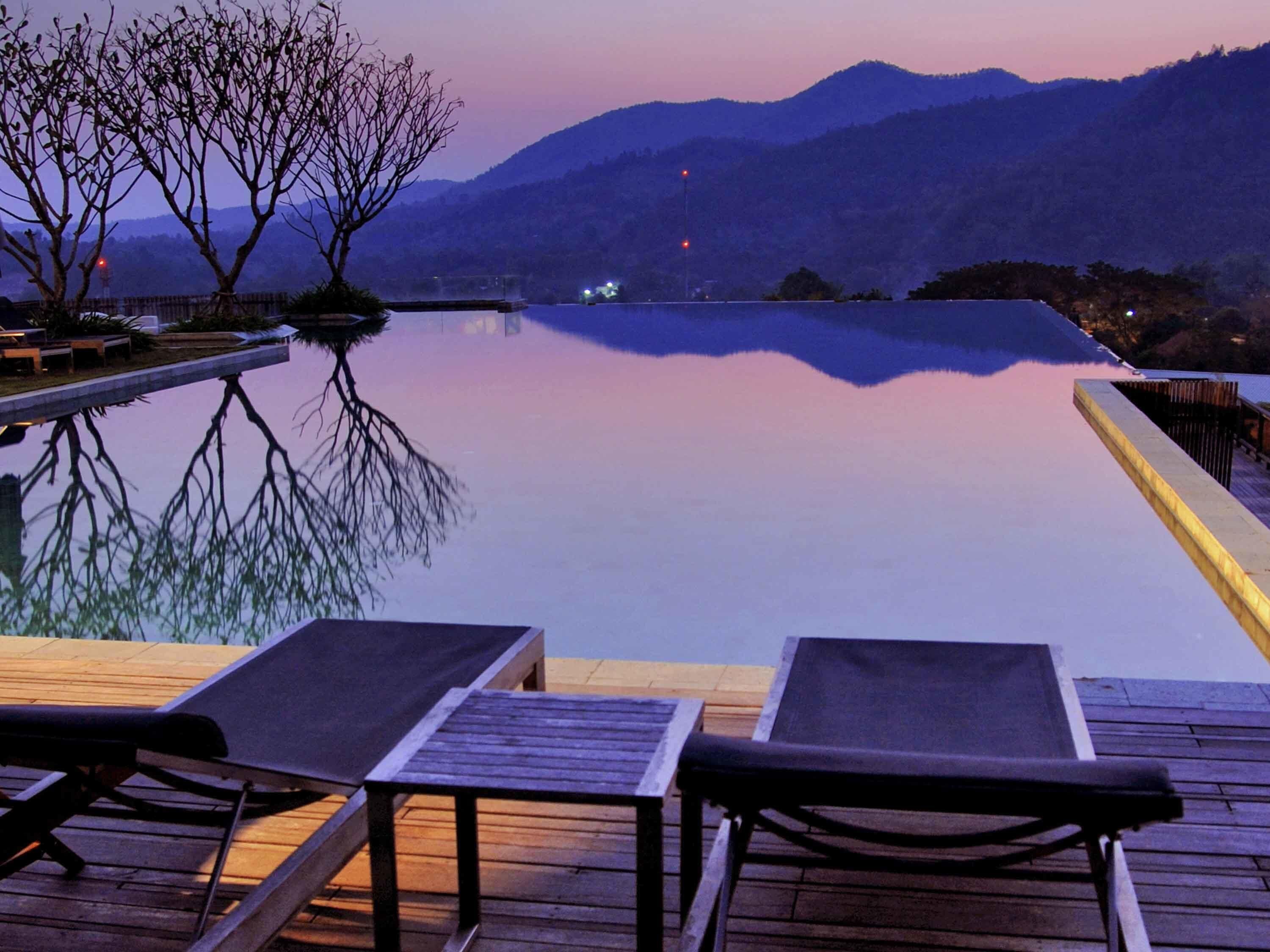 Una piscina infinita con sillones de descanso, una terraza de madera y montañas al fondo.