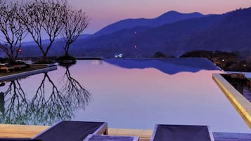 Una piscina infinita con sillones de descanso, una terraza de madera y montañas al fondo.