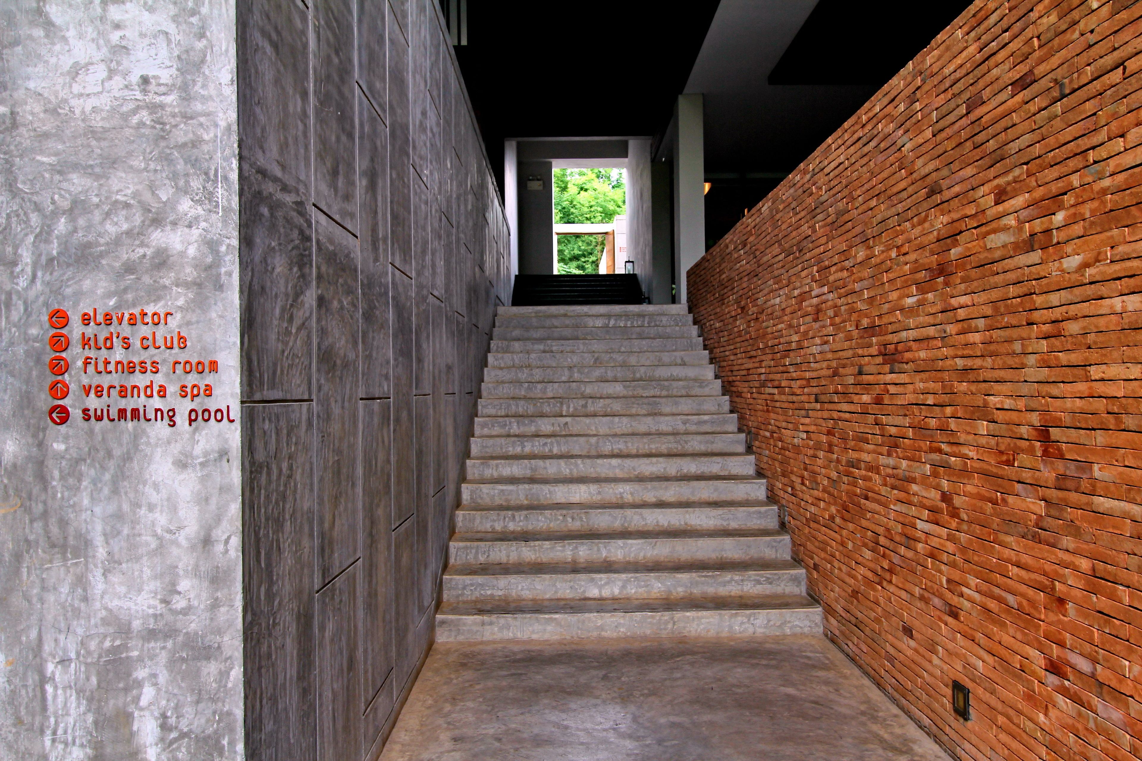 A staircase with a brick wall on one side and a concrete wall on the other, leading to a bright area with greenery visible through a doorway.