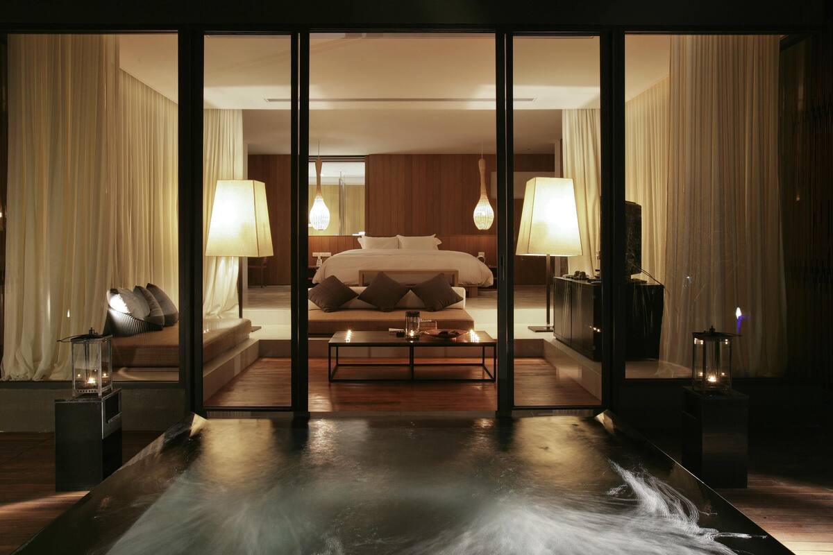 Une chambre d’hôtel moderne avec un grand lit, deux lampes de chevet, une petite table et une vue à travers une paroi en verre.
