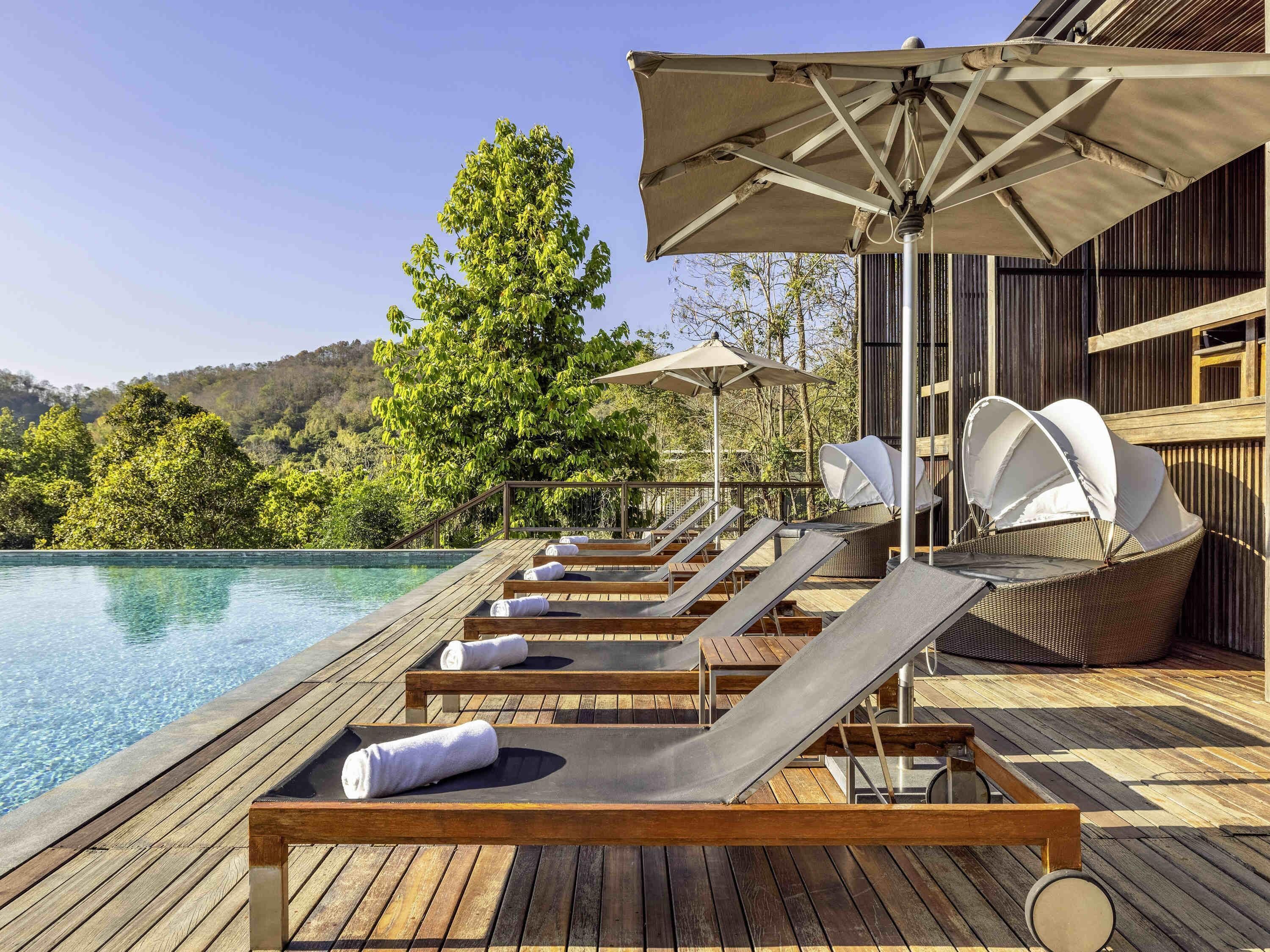 Una terraza de madera con sillones de descanso y sombrillas, con vista a una piscina y una ladera boscosa.