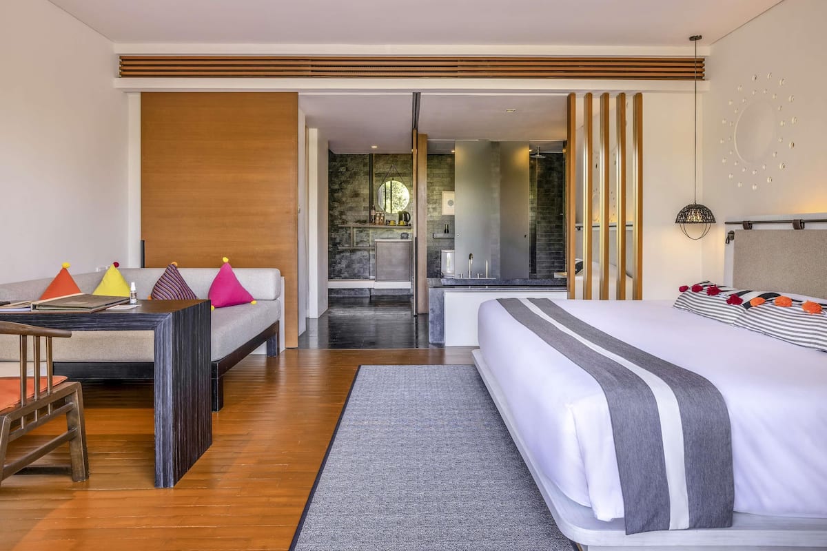 Ein modernes Hotelzimmer mit Bett, Schreibtisch, Sofa und einem durch eine geöffnete Tür einsehbaren Badezimmer.