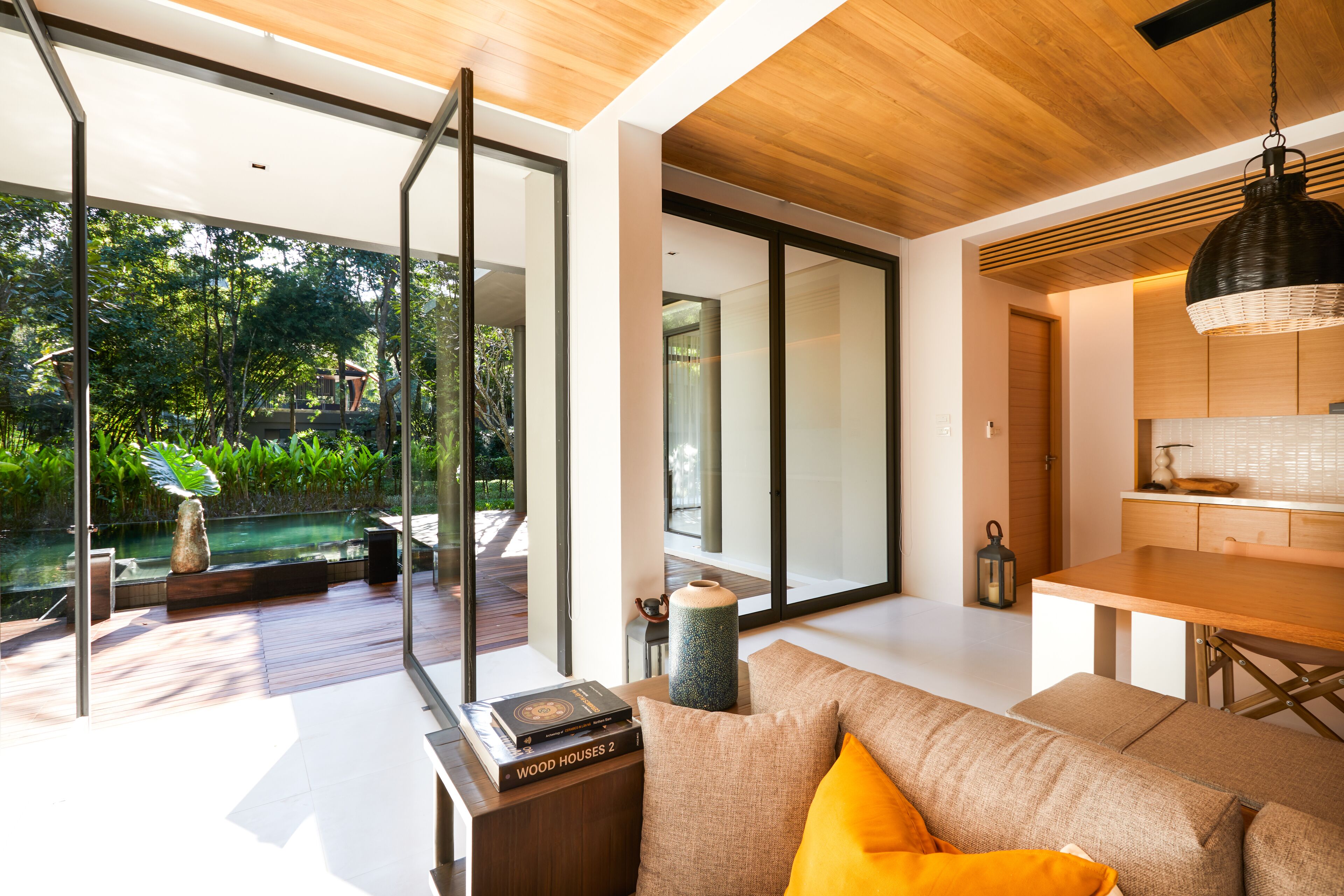 A modern living room with a wooden ceiling, a large window overlooking a garden, a sofa with cushions, and a coffee table with books and a vase.