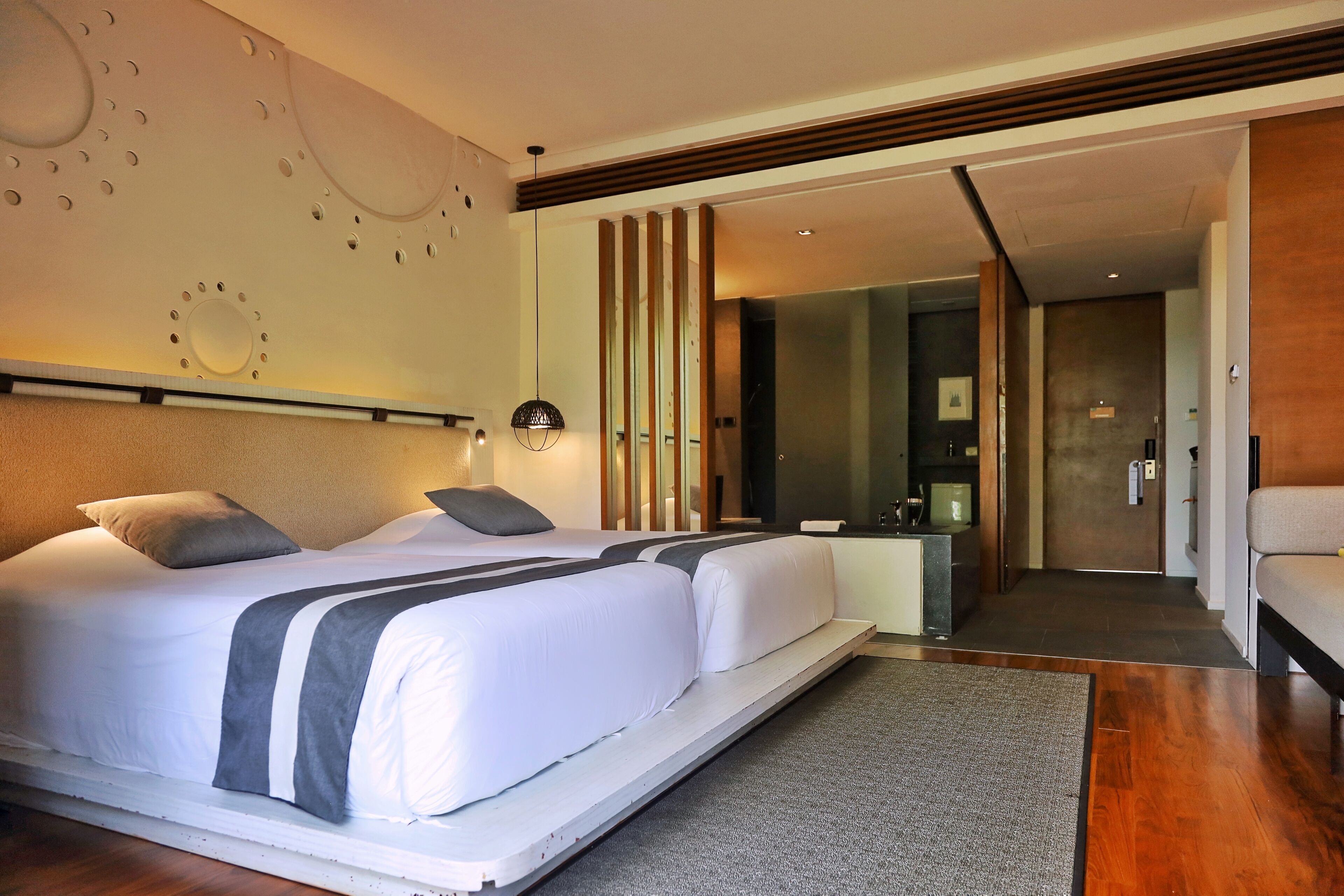 A modern hotel room with a large bed, a bedside table, a mirror, and a view into the hallway.