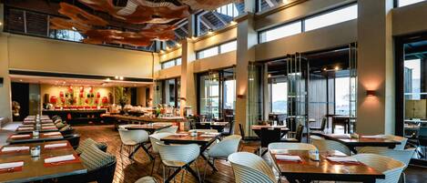 A spacious dining area with high ceilings and large windows. Tables are arranged for groups, and there is a buffet area in the background.