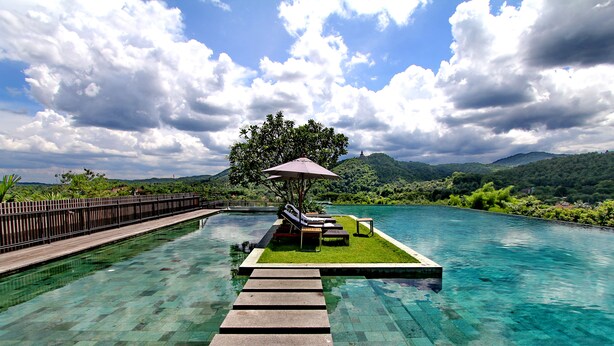 Una piscina cristalina con un deck de madera que conduce a una zona verde con un banco y una sombrilla.