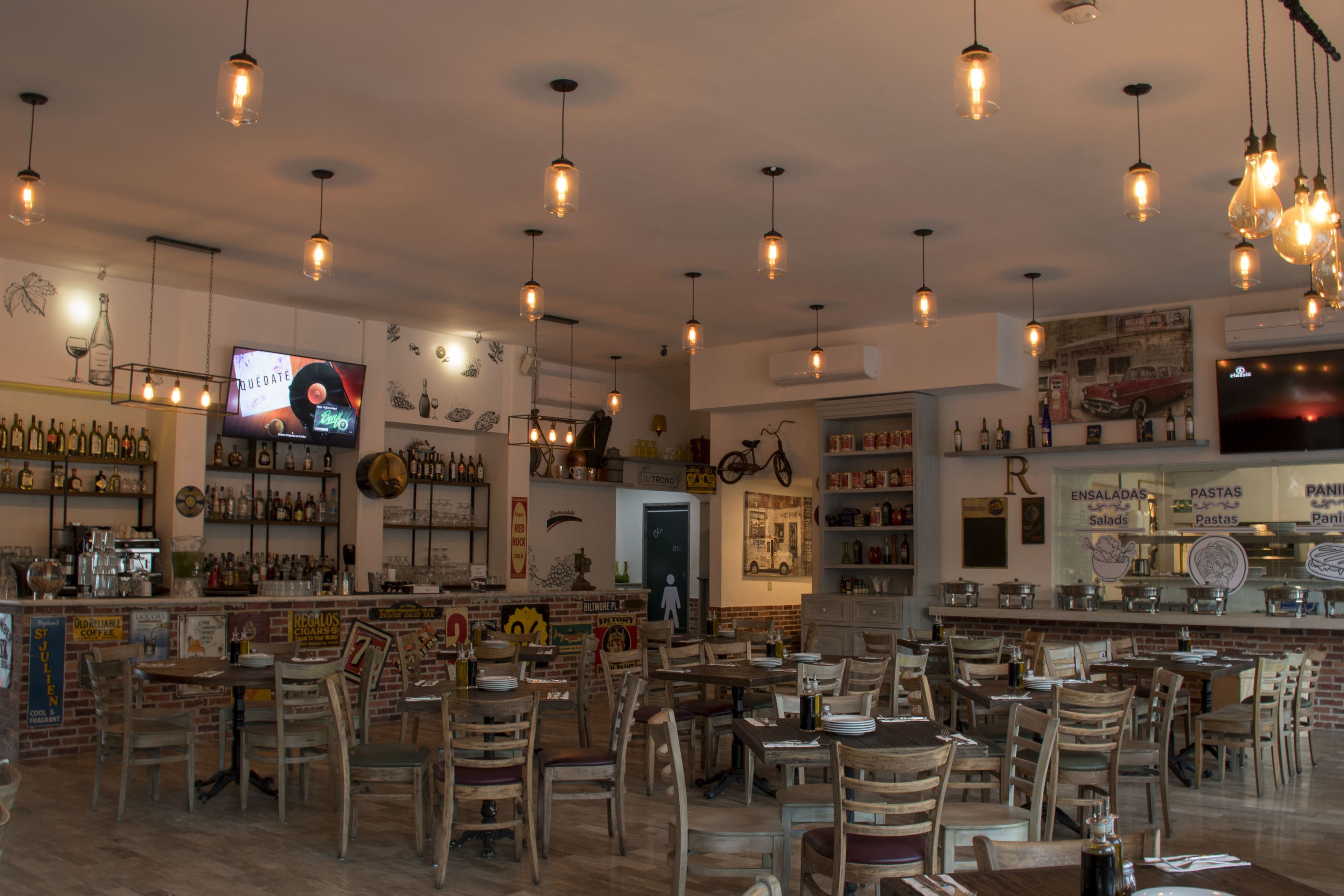 A restaurant interior with wooden tables and chairs, hanging light fixtures, and a bar area with shelves of bottles.