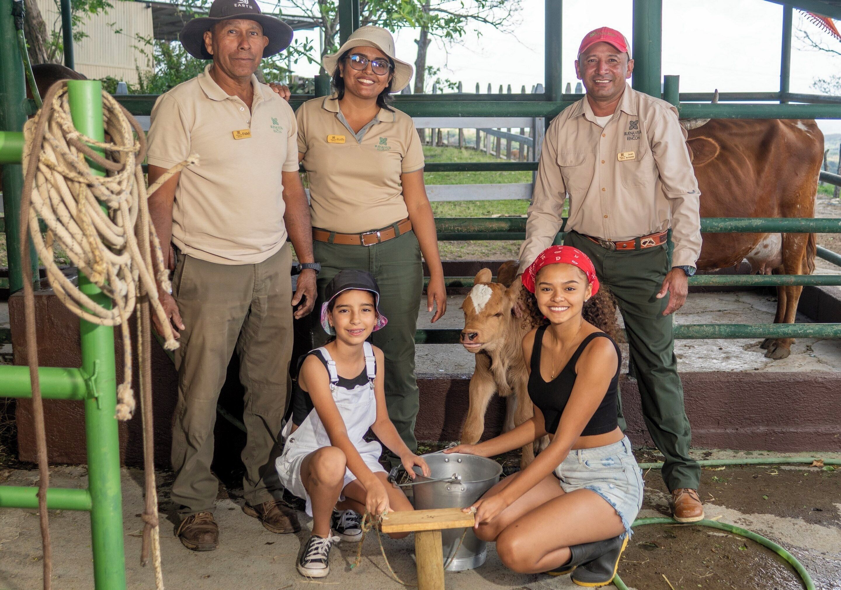Un grupo de personas en un entorno rural, con una vaca y su ternero, de pie cerca de una estructura metálica verde.