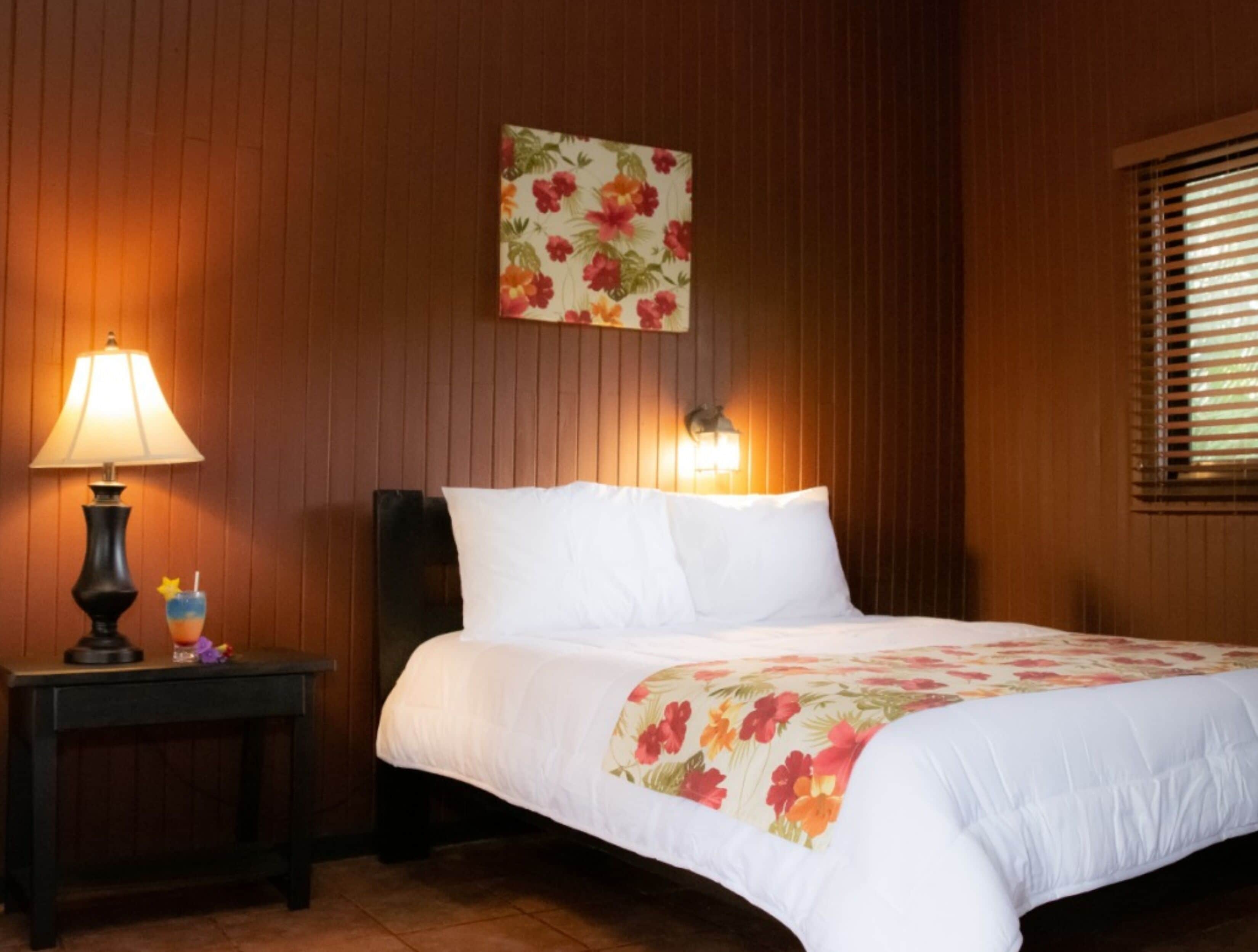 A hotel room with a bed, a nightstand, a lamp, a wall-mounted light, and a window with blinds.