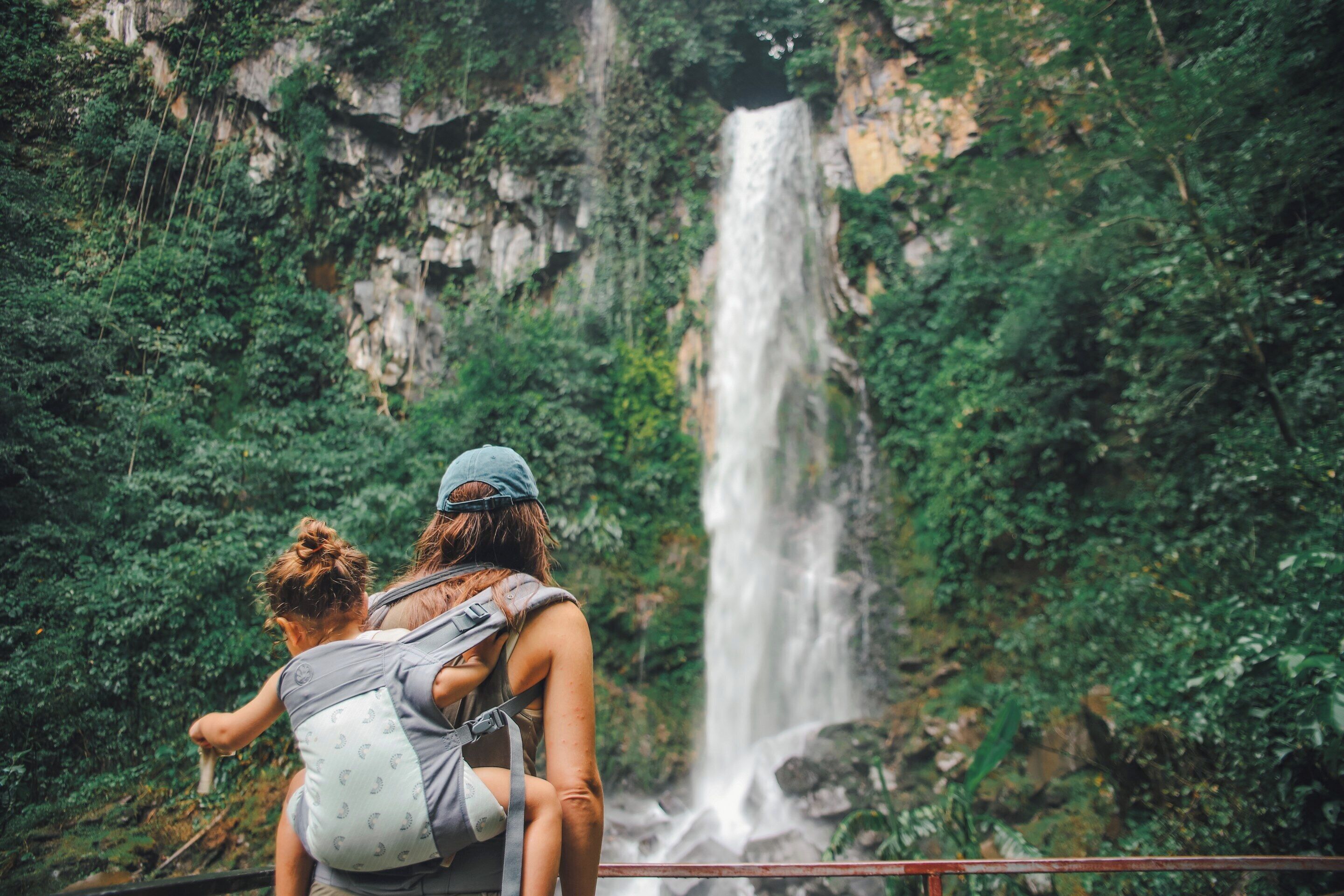 A woman carrying a child on her back, overlooking a waterfall in a lush, green forested area.