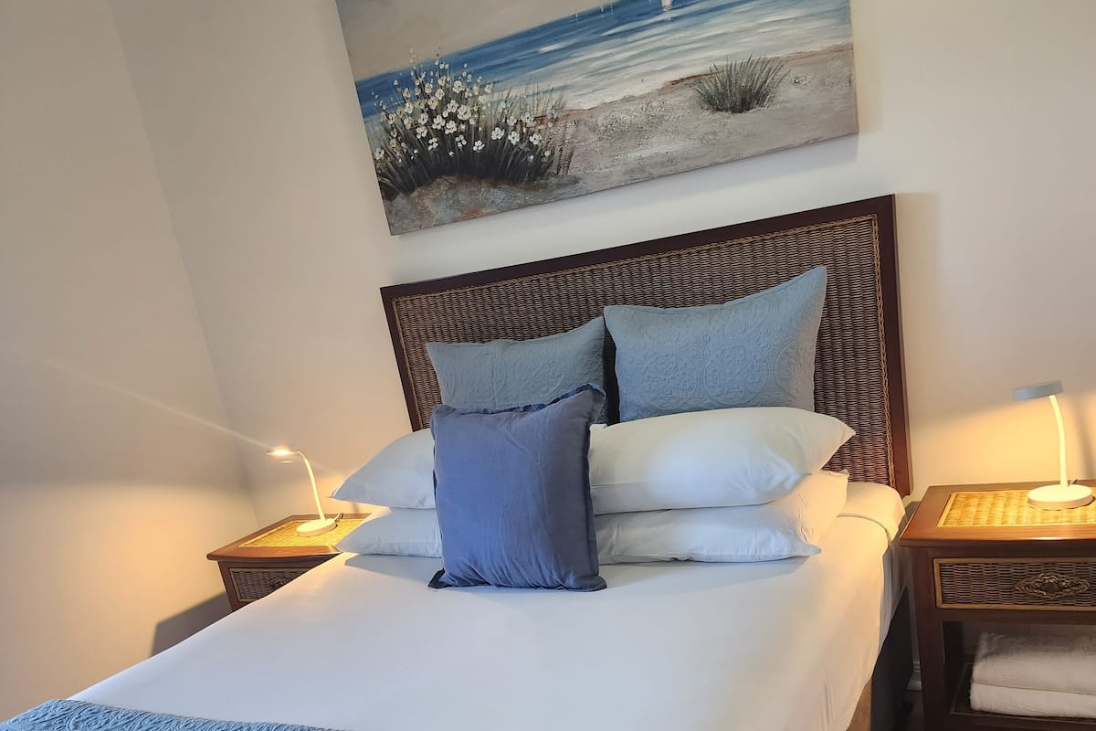 Una cama bien hecha con un edredón azul estampado y almohadas, un cuadro de un paisaje en la pared, y una mesita de noche con una lámpara y una toalla doblada.