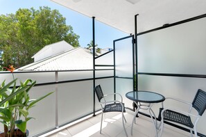 Un balcon avec deux chaises et une table, entouré de rampes de verre, d’où l’on peut admirer des arbres et un bâtiment en arrière-plan.