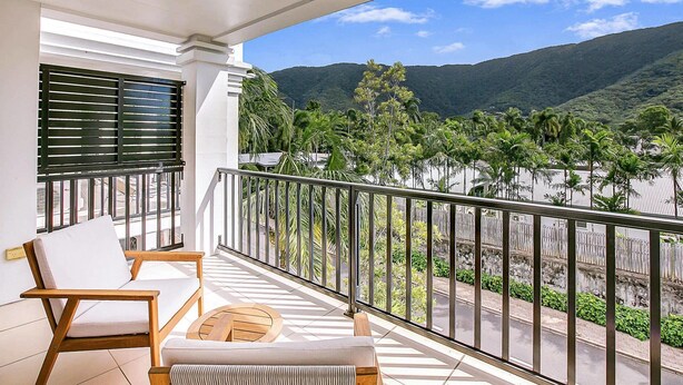 A balcony with a white sofa and a wooden table, overlooking a road and lush green hills. - Elysium Apartments (Palm Cove)