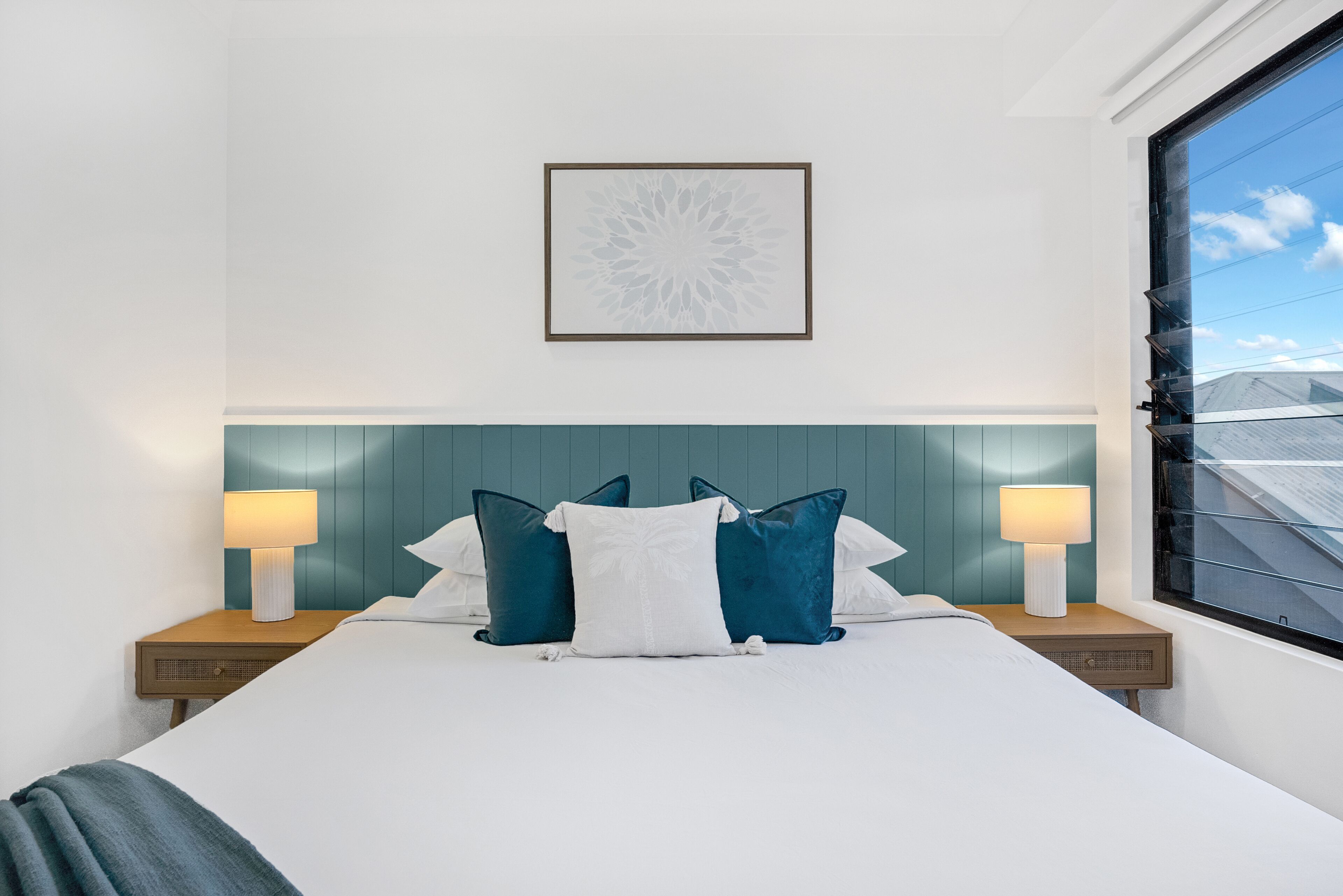 Een net opgemaakt bed met witte en blauwe kussens, twee bedlampjes en een kunstwerk in een lijst aan de muur.