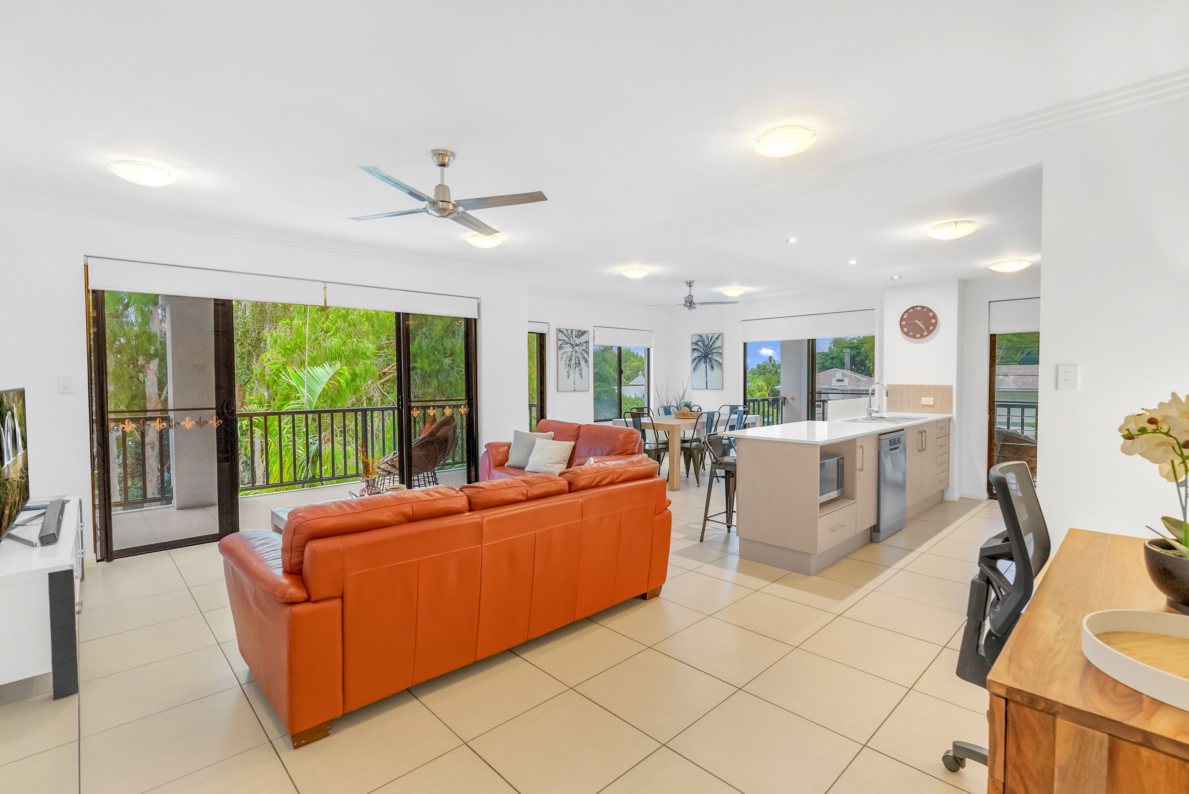 A spacious living area with a large orange sofa, a kitchen with a central island, and large windows offering views of greenery.
