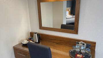 A hotel room with a wooden desk, a mirror, a coffee maker, a tray with bottled water and snacks, and a bed with a dark headboard.