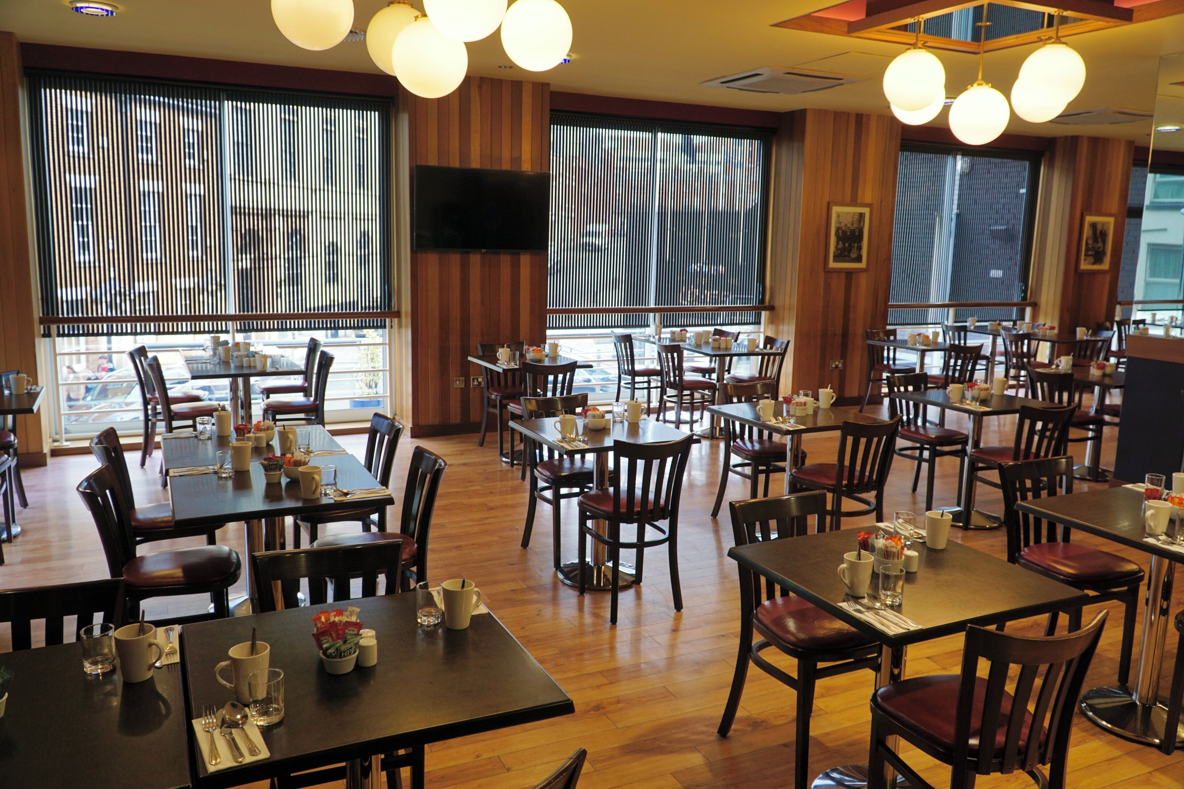 A restaurant with multiple tables and chairs, wooden floors, and large windows with blinds.