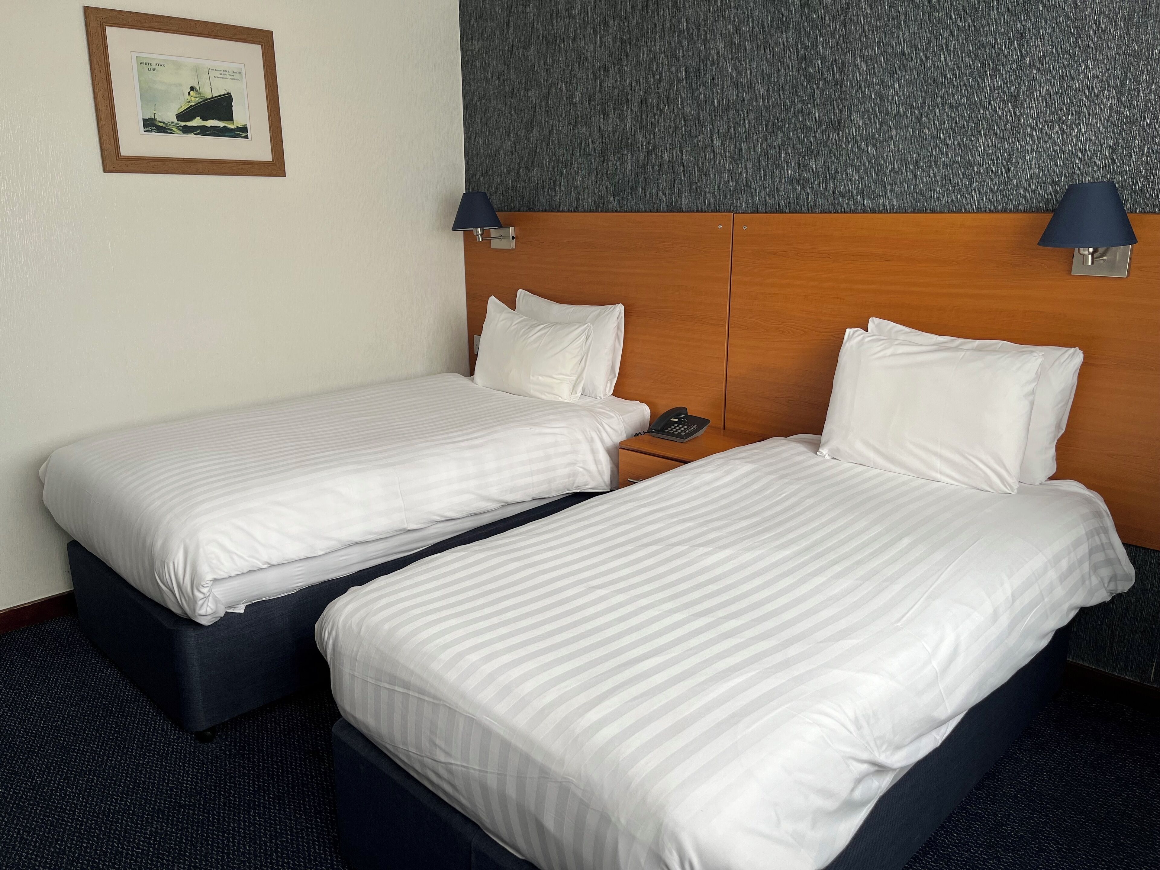 Ett hotellrum med två separata sängar, en sänggavel i trä, en inramad tavla på väggen och två vägglampor.