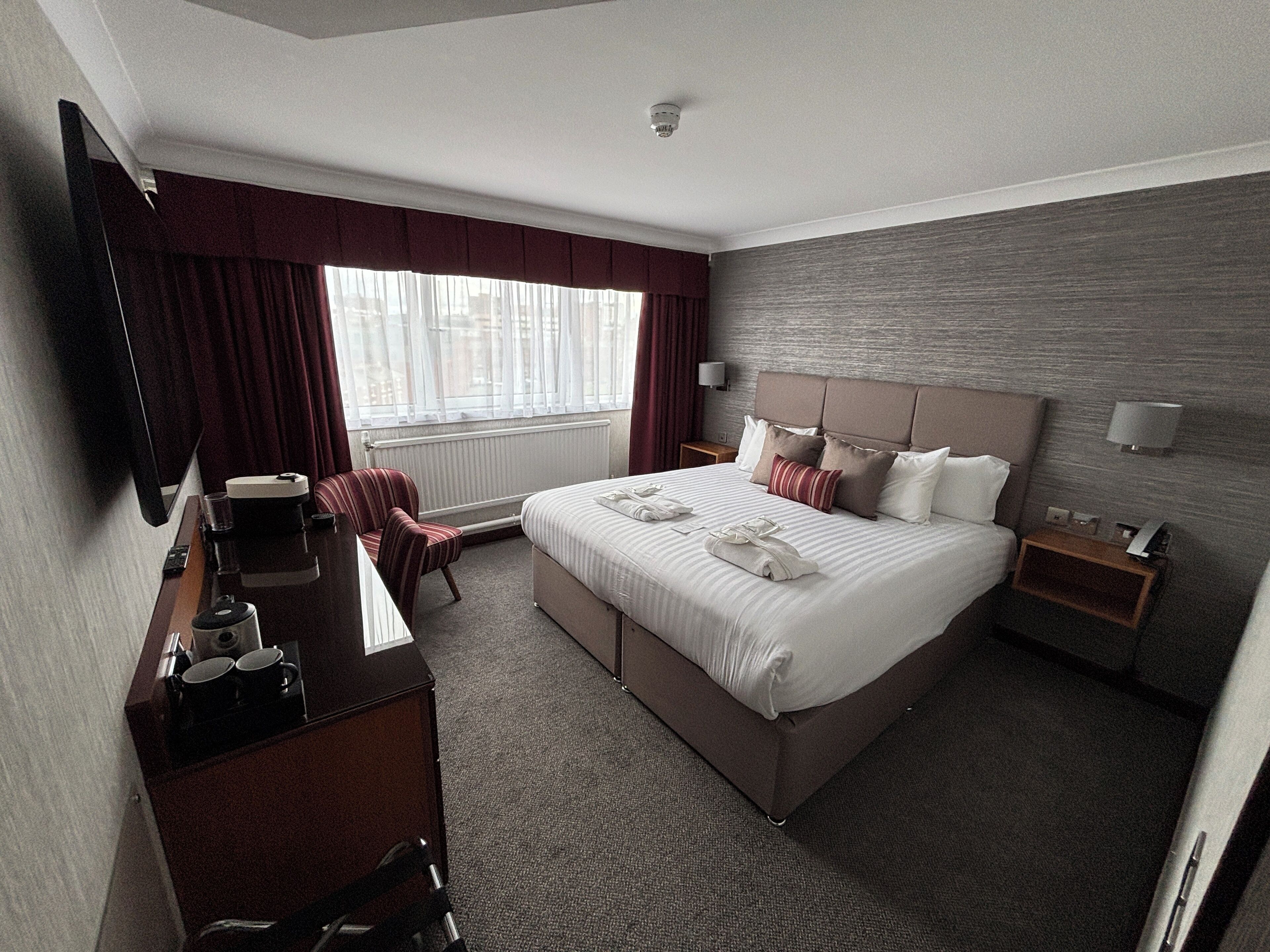 A hotel room with a large bed, a desk with a chair, a bedside table, and a window with curtains.