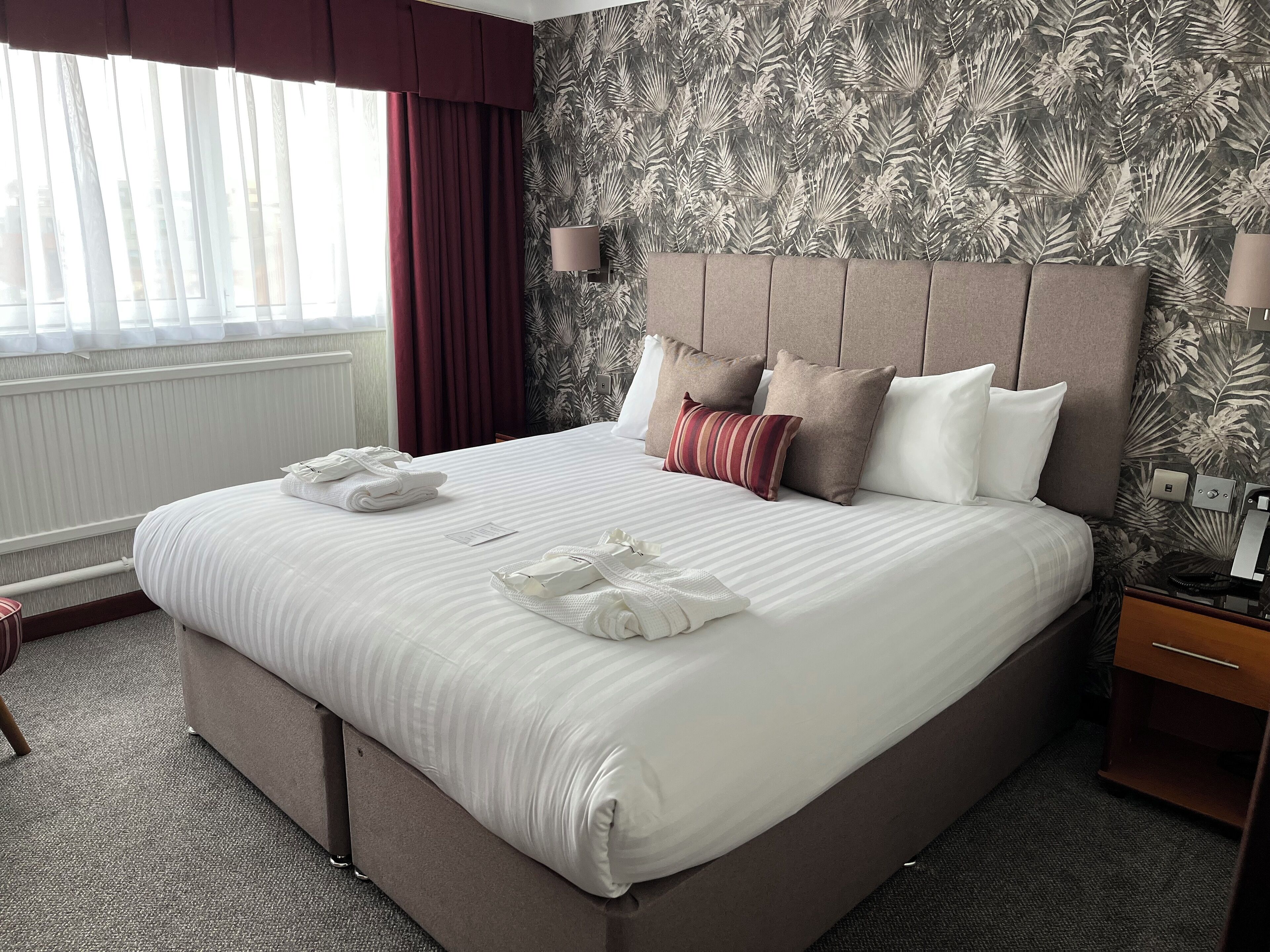 Een netjes opgemaakt bed met witte lakens en een gestreept kussen, een nachtkastje met een lamp, en een raam met gordijnen.