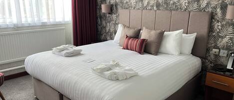 Een netjes opgemaakt bed met witte lakens en een gestreept kussen, een nachtkastje met een lamp, en een raam met gordijnen.