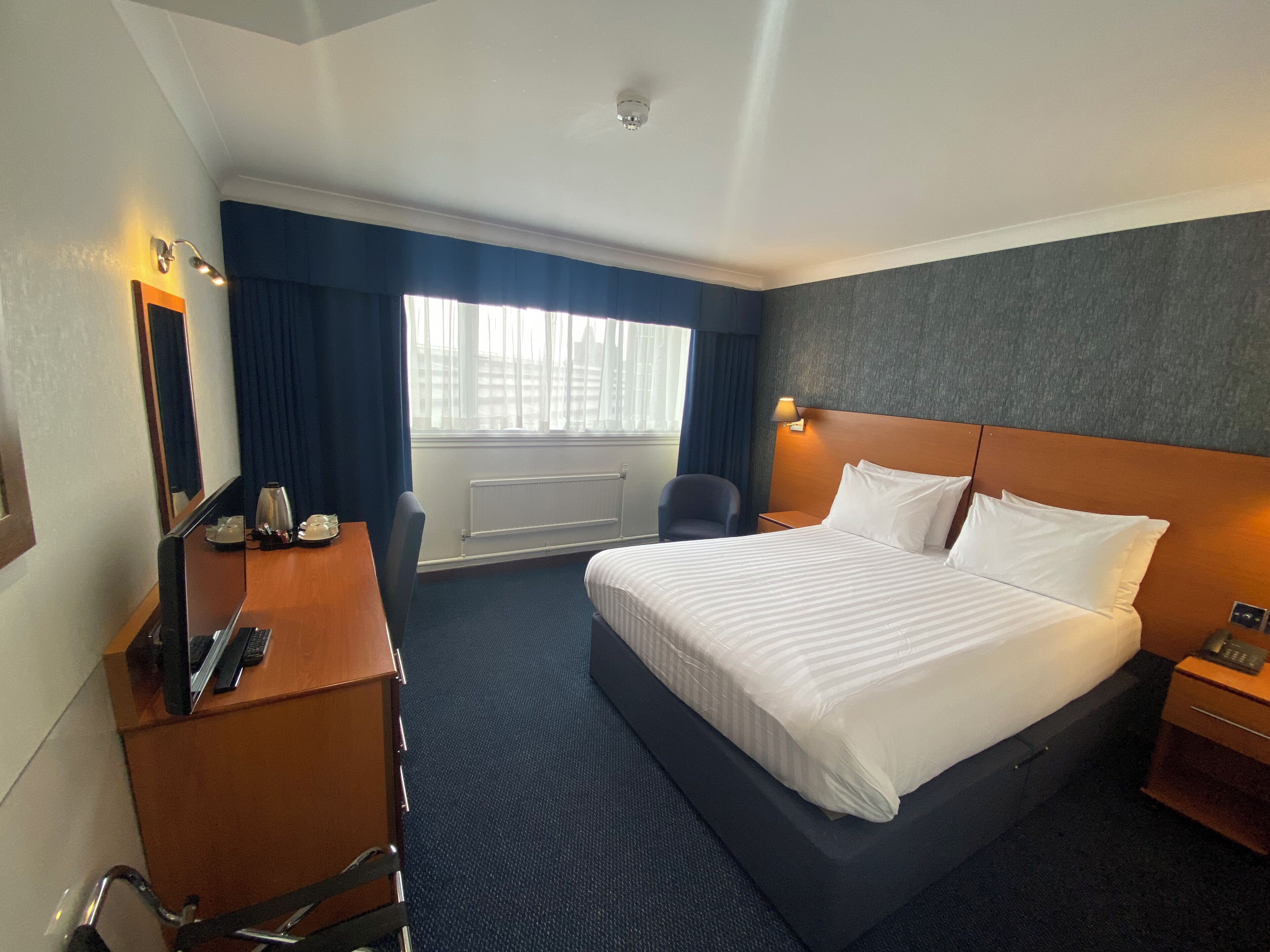 Habitación de hotel con una cama grande, un escritorio con ordenador, una silla y una ventana con cortinas.