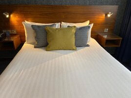 Una cama bien hecha con sábanas blancas y tres almohadas, dos estampadas y una verde lisa, en una habitación con cabecera y mesitas de noche de madera.