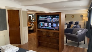 Camera d'albergo con una grande TV a schermo piatto montata su un supporto in legno, un divano, una scrivania con una sedia e un bagno parzialmente visibile attraverso una porta aperta.