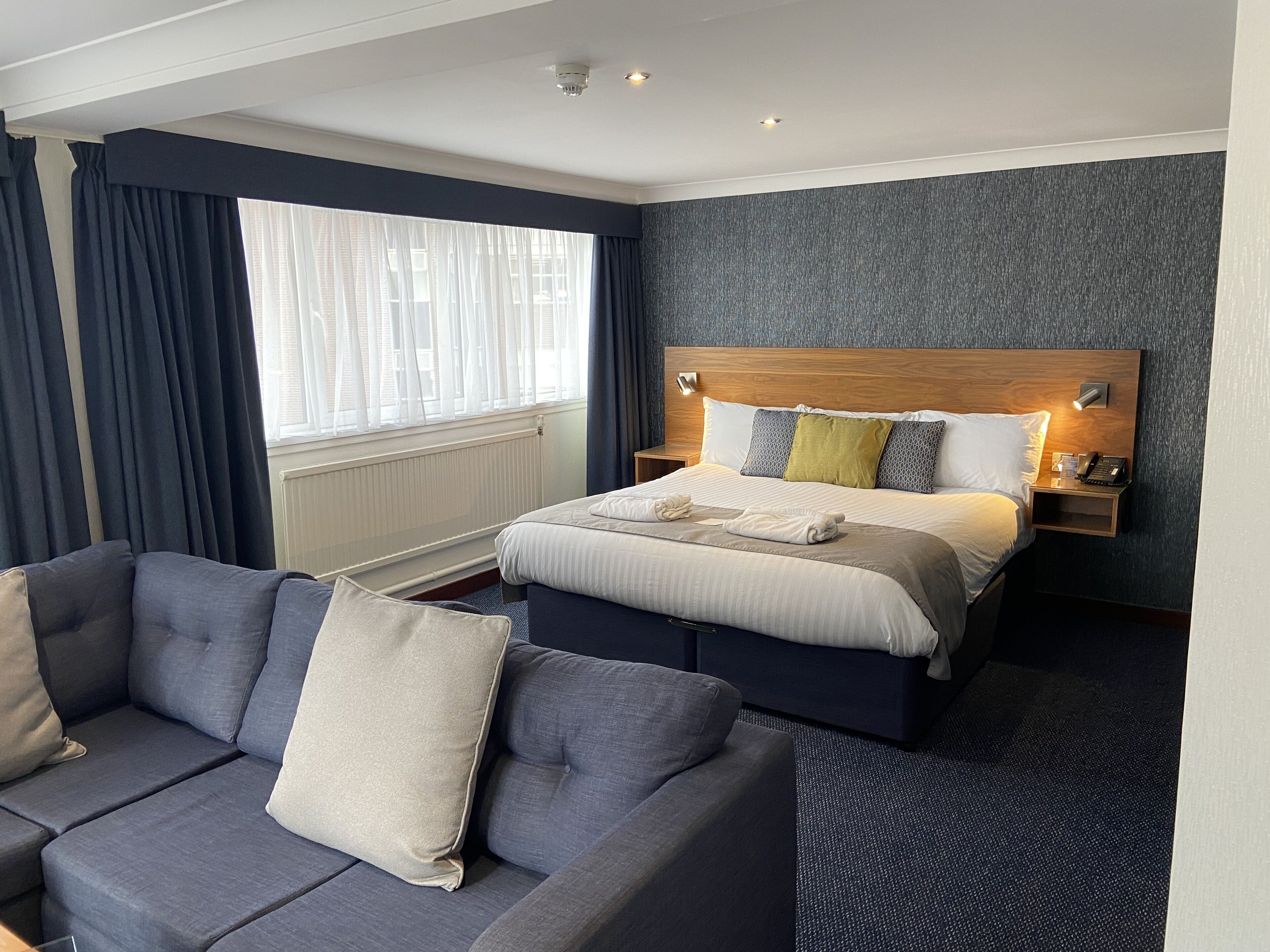 Una habitación de hotel con cama, sofá, mesita de noche y ventana con cortinas.