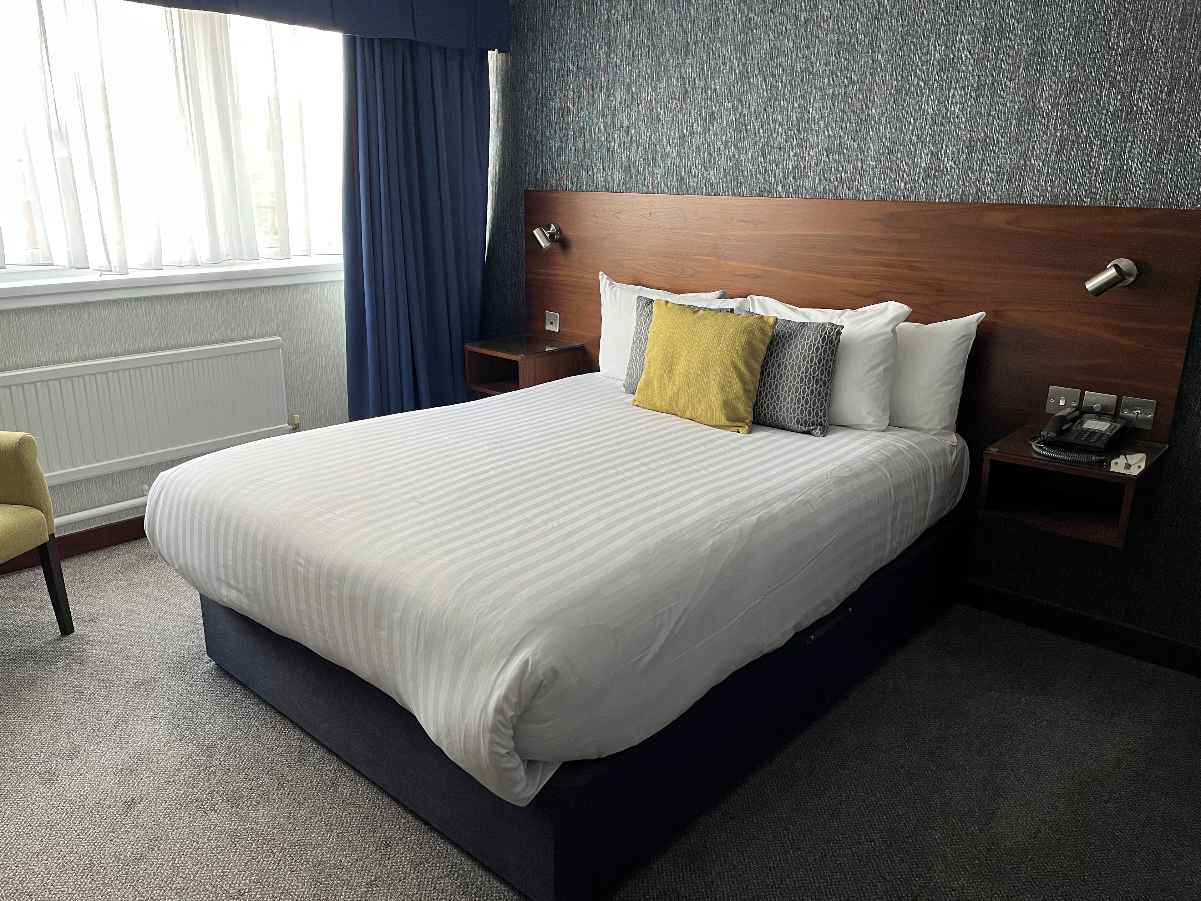 A hotel room with a large bed, a bedside table with a phone, a nightstand, a chair, and a window with curtains.