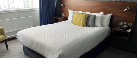 Une chambre d’hôtel avec un grand lit, une table de chevet avec un téléphone, une commode, une chaise et une fenêtre avec des rideaux.