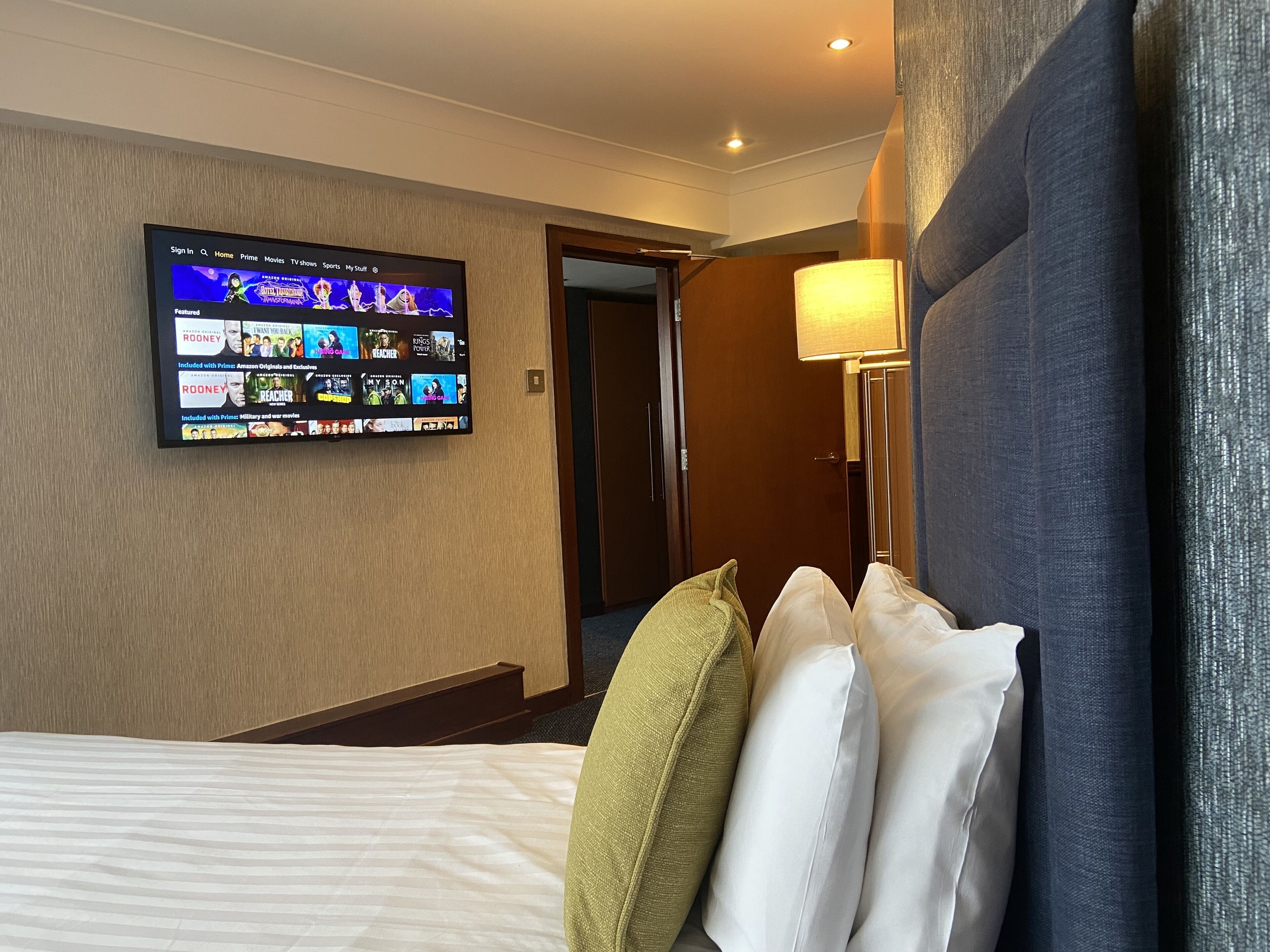 A hotel room with a large flat-screen TV mounted on the wall displaying a streaming service interface. There is a bed with white and green pillows, a bedside lamp, and a wooden door leading to another room.