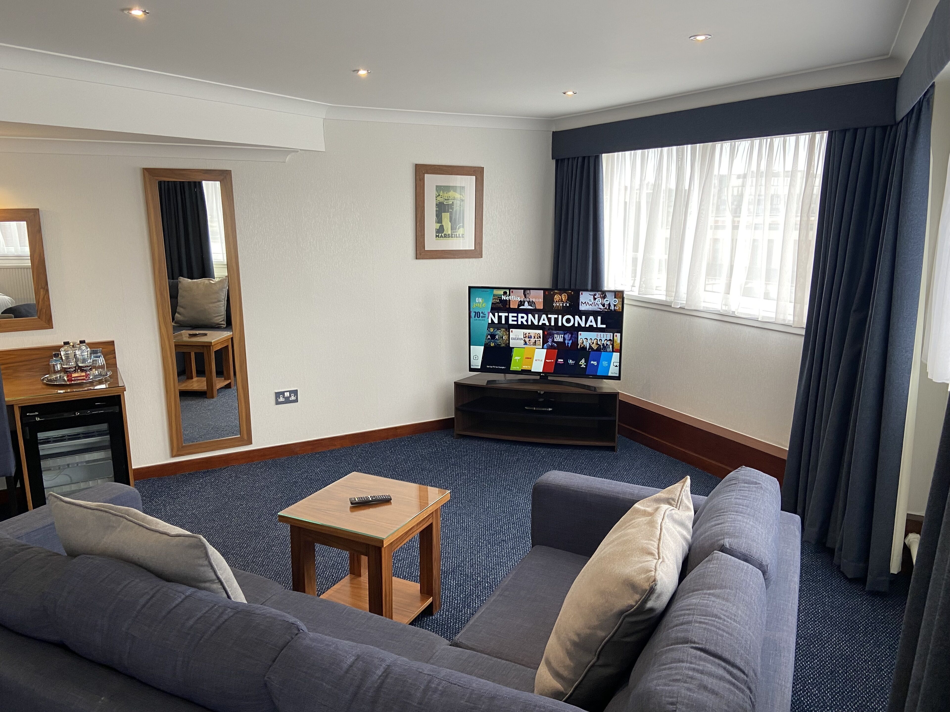Camera d'albergo con una grande televisione a schermo piatto che mostra una guida ai canali internazionali, un divano grigio con cuscini, un piccolo tavolino in legno e un'ampia finestra con tende.