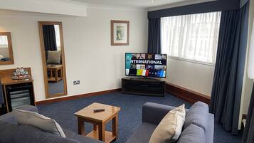Camera d'albergo con una grande televisione a schermo piatto che mostra una guida ai canali internazionali, un divano grigio con cuscini, un piccolo tavolino in legno e un'ampia finestra con tende.