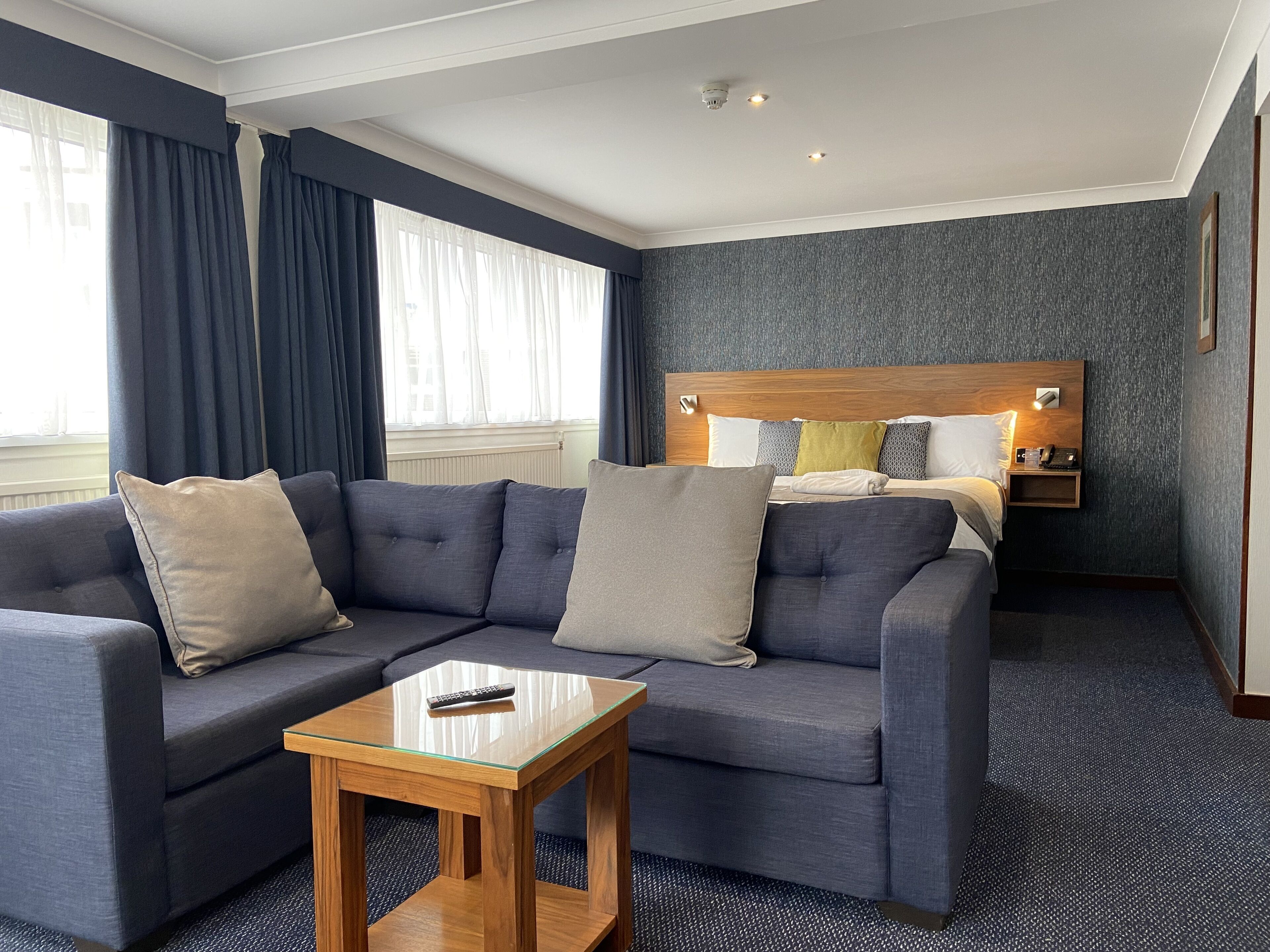 A modern hotel room with a grey sofa, a wooden side table, a bed with a wooden headboard, and a grey textured wall.