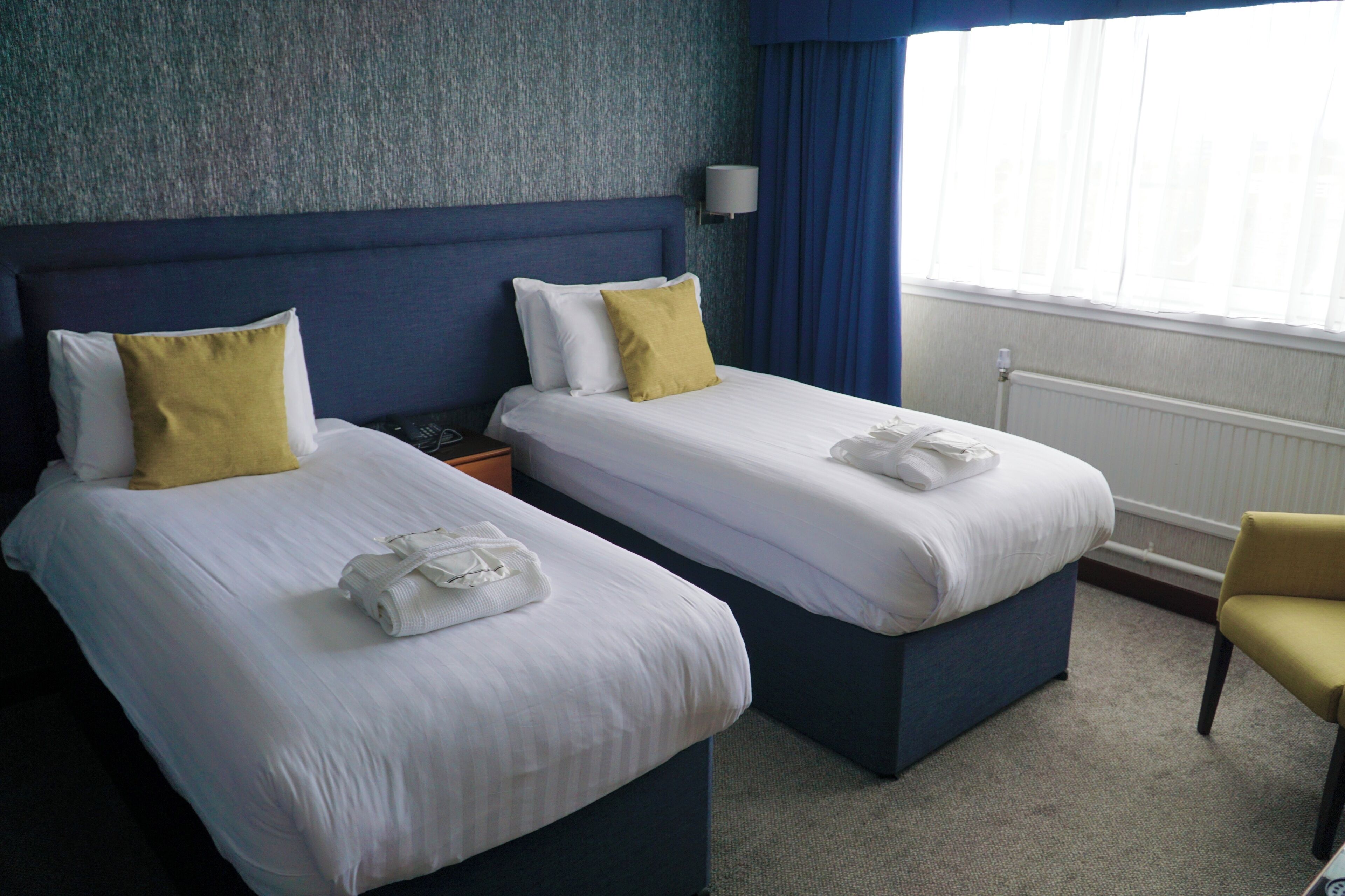 A hotel room with two beds, a blue headboard, a white wall, a window with blue curtains, and a chair.