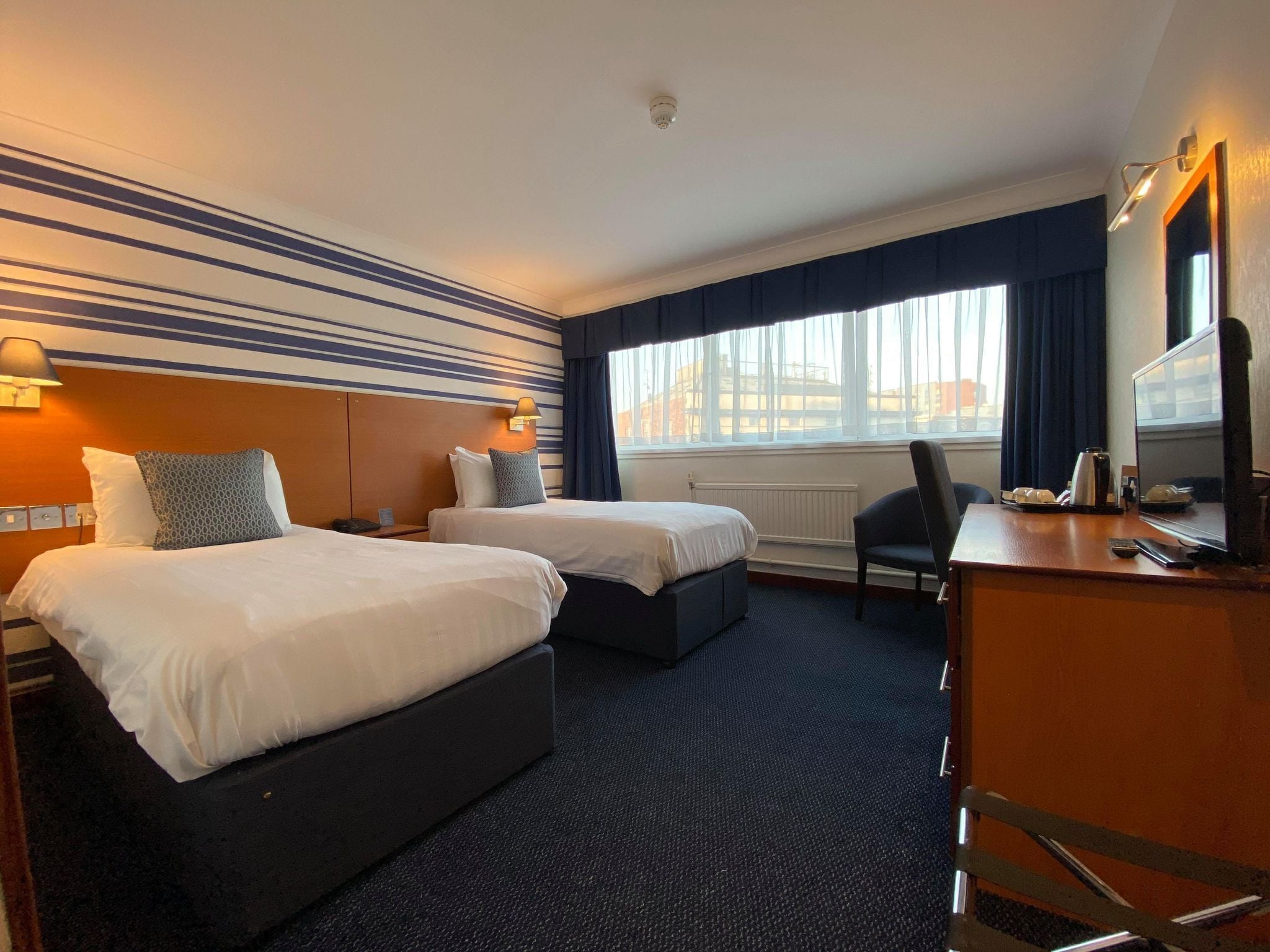 A hotel room with two beds, a desk, a chair, a TV, and a window with curtains.