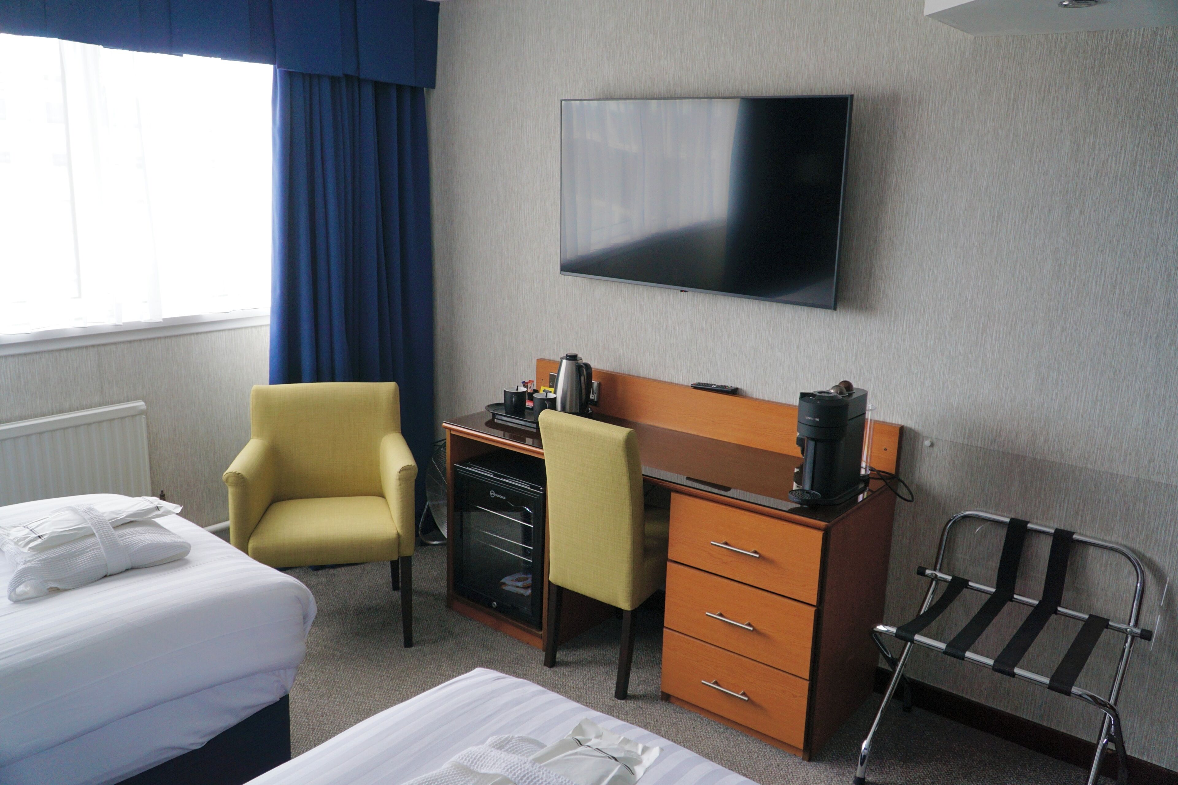 Una habitación de hotel con una cama, un escritorio con computadora, una silla, un televisor montado en la pared y una ventana con cortinas.
