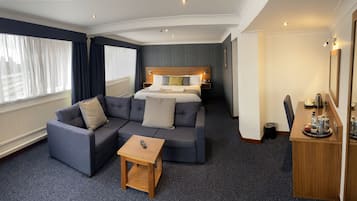 A hotel room with a sofa set, a small table, a bed with a wooden headboard, and a bedside table with a tray of amenities.