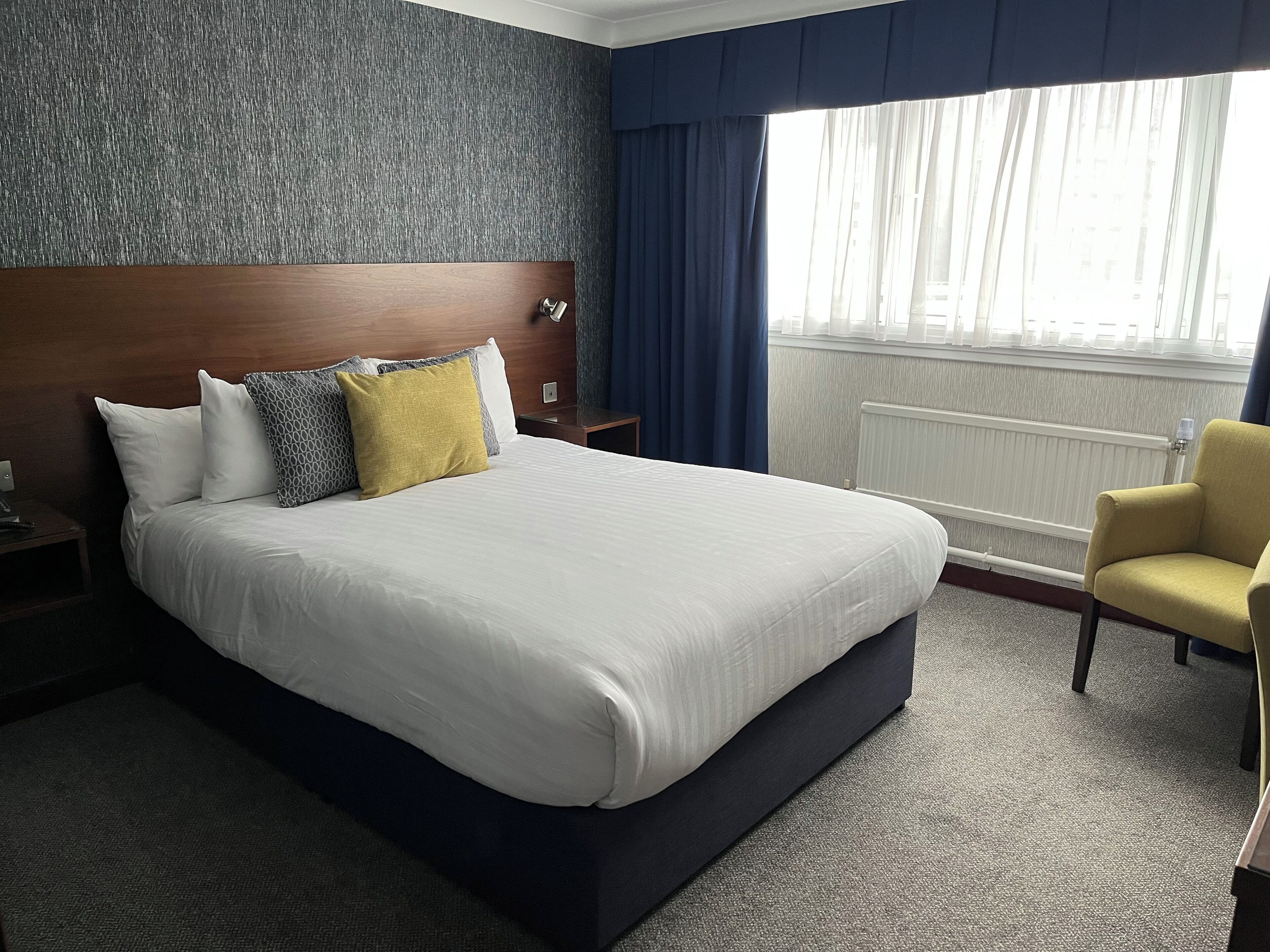 A hotel room with a large bed, a wooden headboard, a nightstand, a chair, and a window with curtains.
